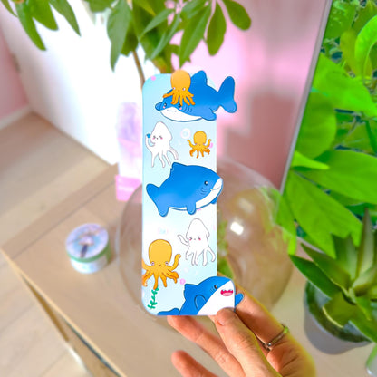 Hand holding the Pastel Shark Bookmark in a cozy workspace with a pink wall and leafy green plant in the background. The ocean animal page marker shows chibi sharks, octopus, and squid swimming through soft blue water. This lifestyle photo demonstrates real-life scale, thickness, and how the double-sided matte laminated bookmark looks in hand for readers and stationery lovers.