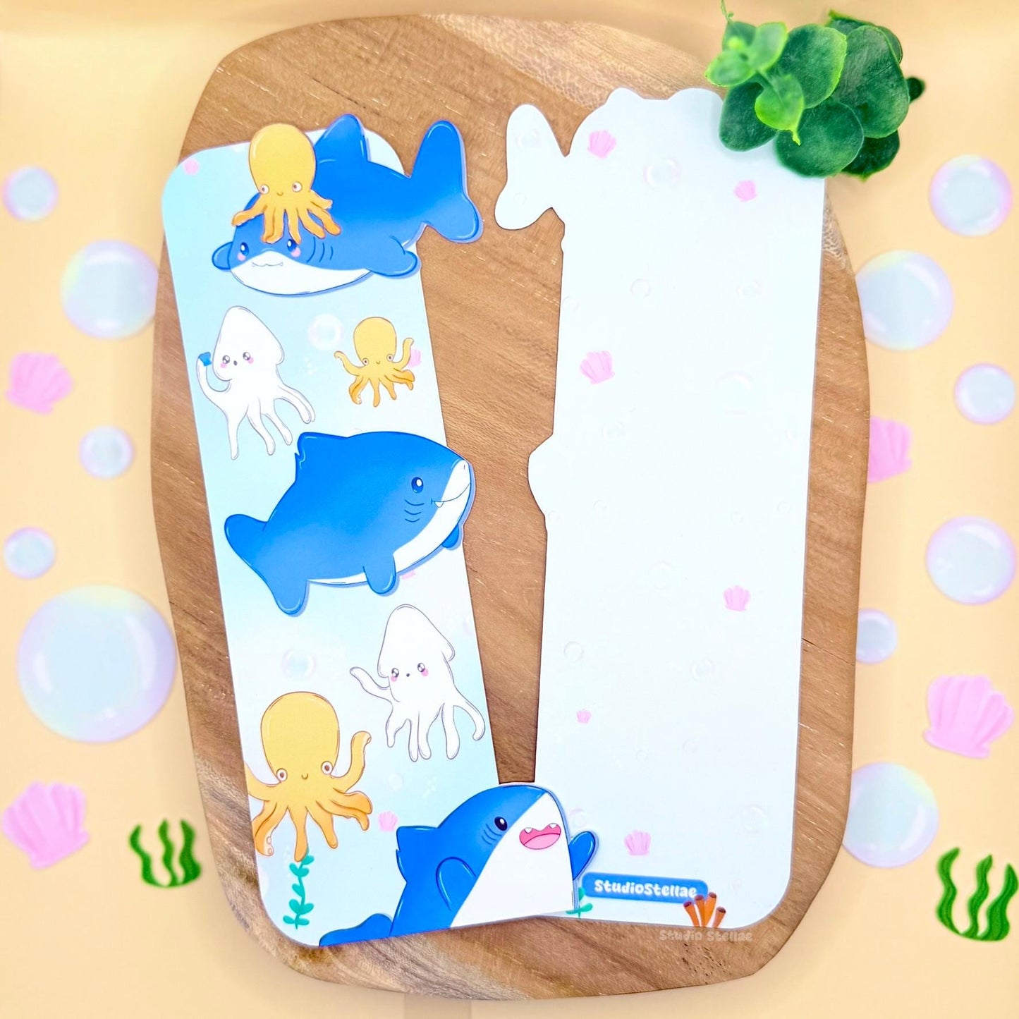 Pastel Shark Bookmark displayed upright on a warm wooden surface with a soft yellow polka dot background. The uniquely shaped ocean animal page marker features chibi blue sharks, orange octopus, and cute squid floating in pastel blue water. The matte laminated finish and rounded edges are visible, highlighting the kawaii sea animals design and cheerful underwater theme perfect for book lovers and shark fans.