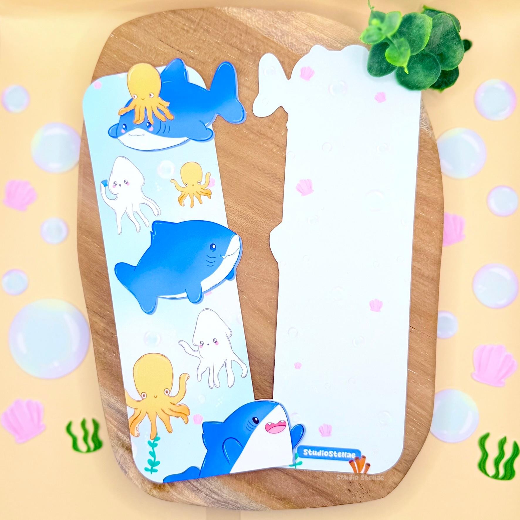 Pastel Shark Bookmark displayed upright on a warm wooden surface with a soft yellow polka dot background. The uniquely shaped ocean animal page marker features chibi blue sharks, orange octopus, and cute squid floating in pastel blue water. The matte laminated finish and rounded edges are visible, highlighting the kawaii sea animals design and cheerful underwater theme perfect for book lovers and shark fans.