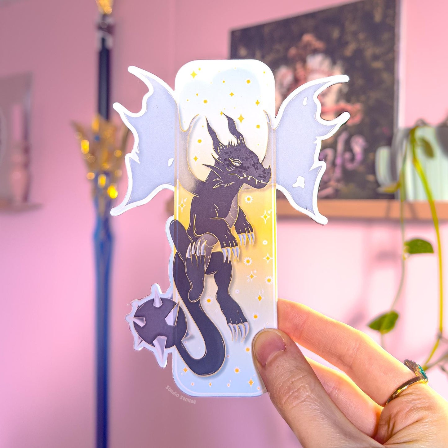 Close-up of a hand holding the pastel fantasy dragon bookmark, showing the front design with a dark blue dragon silhouette, starry details, and golden clouds. The die-cut dragon wings extend beyond the bookmark edges, making the shape look bold and unique. The smooth matte lamination and sturdy thickness are visible, presenting this Fourth Wing inspired romantasy page marker as a BookTok reader gift for dragon lovers.