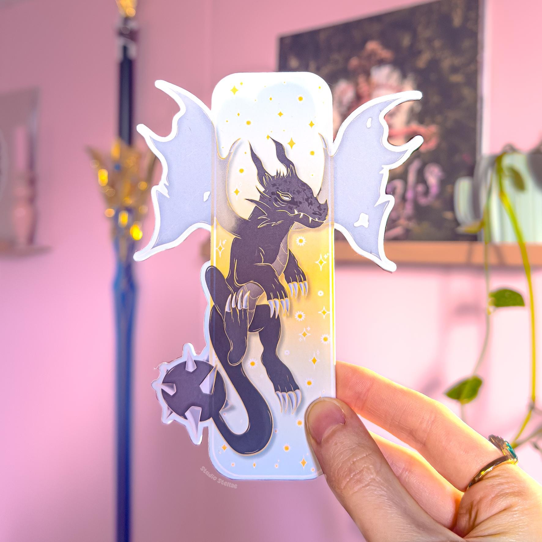 Close-up of a hand holding the pastel fantasy dragon bookmark, showing the front design with a dark blue dragon silhouette, starry details, and golden clouds. The die-cut dragon wings extend beyond the bookmark edges, making the shape look bold and unique. The smooth matte lamination and sturdy thickness are visible, presenting this Fourth Wing inspired romantasy page marker as a BookTok reader gift for dragon lovers.