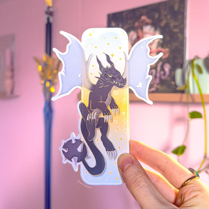 Close-up of a hand holding the pastel fantasy dragon bookmark, showing the front design with a dark blue dragon silhouette, starry details, and golden clouds. The die-cut dragon wings extend beyond the bookmark edges, making the shape look bold and unique. The smooth matte lamination and sturdy thickness are visible, presenting this Fourth Wing inspired romantasy page marker as a BookTok reader gift for dragon lovers.