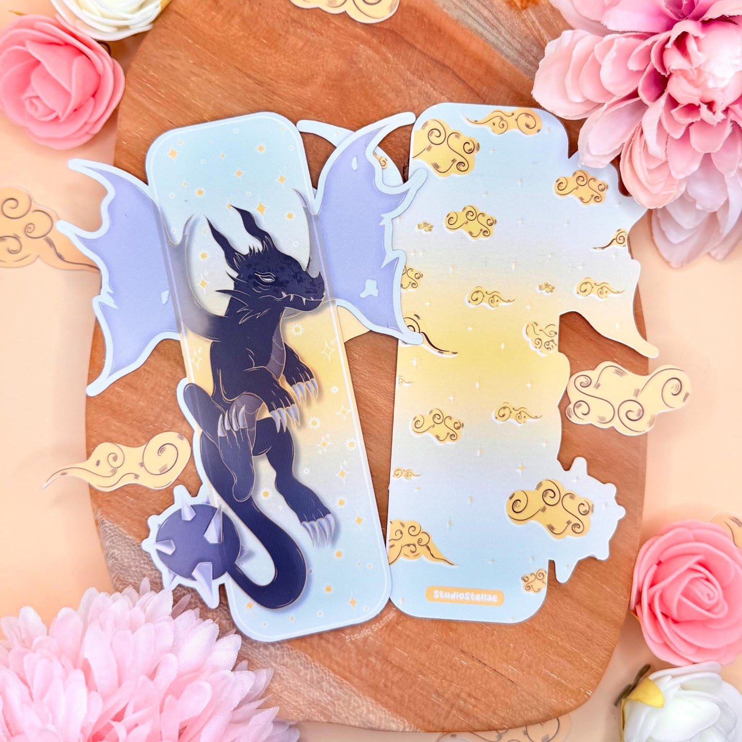 Pastel fantasy dragon bookmark styled on a warm wooden board with pink flowers and golden cloud accents around it. The uniquely shaped laminated page marker shows a dark blue shadow dragon with wide wings and golden stars on a soft pastel sky background. Dreamy romantasy aesthetic inspired by Fourth Wing and the Empyrean series, perfect as a BookTok reader gift for dragon lovers and fantasy book fans. Matte laminated finish and die-cut shape are clearly visible.