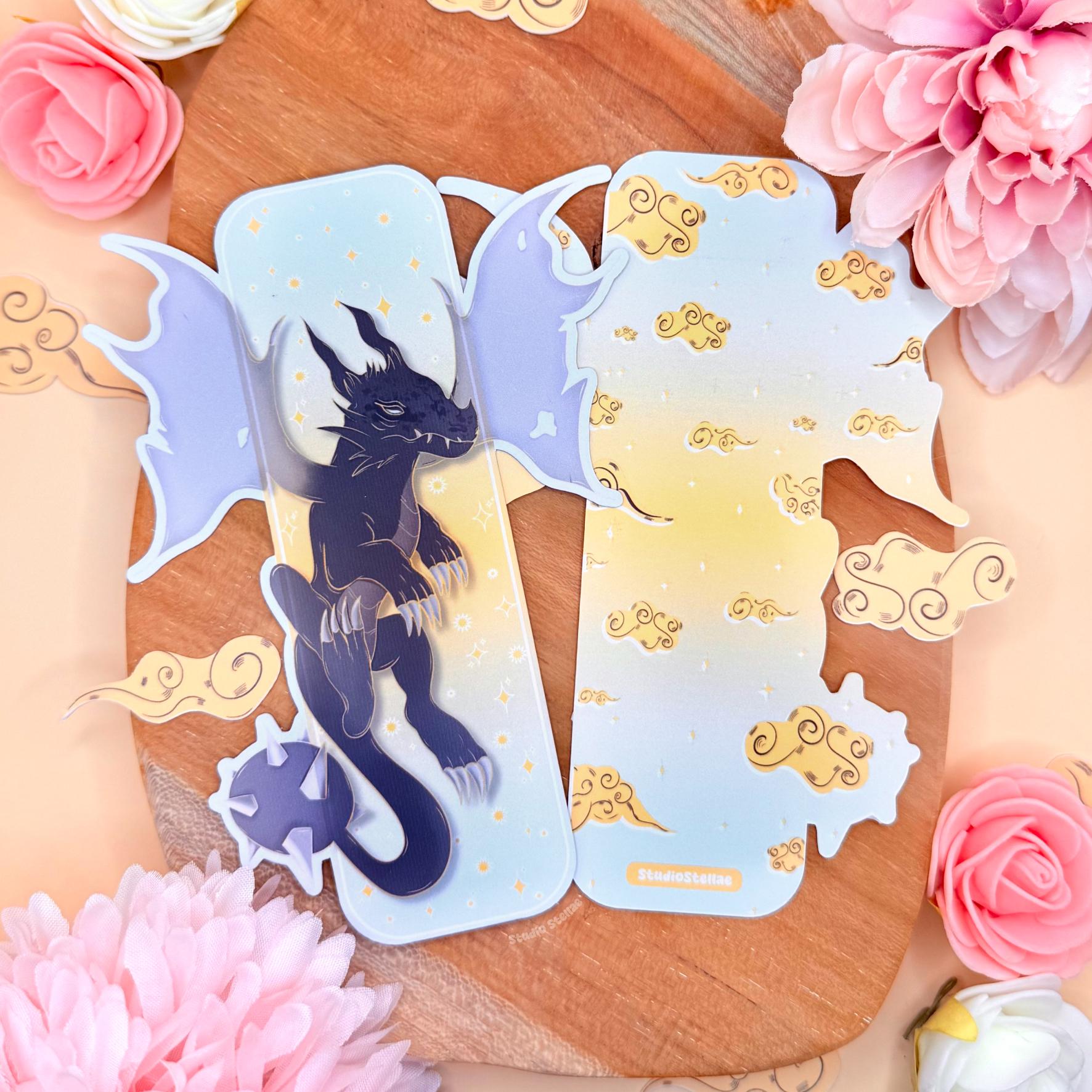 Pastel fantasy dragon bookmark styled on a warm wooden board with pink flowers and golden cloud accents around it. The uniquely shaped laminated page marker shows a dark blue shadow dragon with wide wings and golden stars on a soft pastel sky background. Dreamy romantasy aesthetic inspired by Fourth Wing and the Empyrean series, perfect as a BookTok reader gift for dragon lovers and fantasy book fans. Matte laminated finish and die-cut shape are clearly visible.