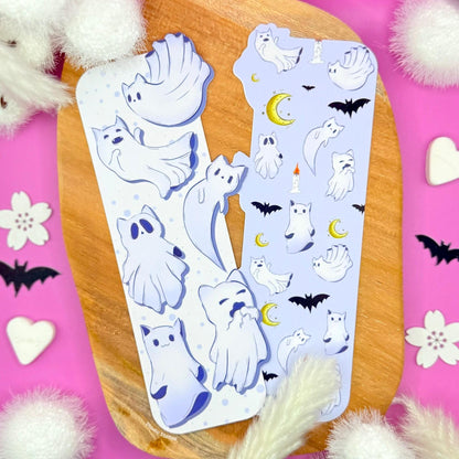 Witchy Cat Ghost Bookmark displayed on a wooden board surrounded by pink heart decorations and soft white pom details. The uniquely shaped die-cut bookmark features floating ghost cats in pastel purple tones with playful expressions, moons, and spooky Halloween accents. This double-sided ghost cat bookmark highlights the whimsical design and laminated finish, perfect as a spooky reader gift or Halloween kitty bookmark for book lovers.