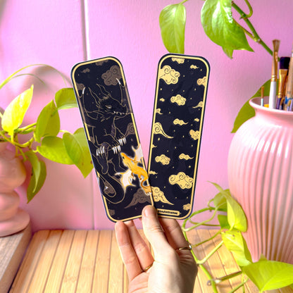 Hand holding two dark fantasy dragon bookmarks in a cozy workspace with a pink wall, leafy green plant, and pastel decor in the background. The matte laminated finish and slim rectangular shape are clearly visible. The design showcases a black shadow dragon and a glowing golden dragon against a deep midnight sky, demonstrating real-life scale and how the romantasy page marker looks when used by BookTok readers and fantasy fans.