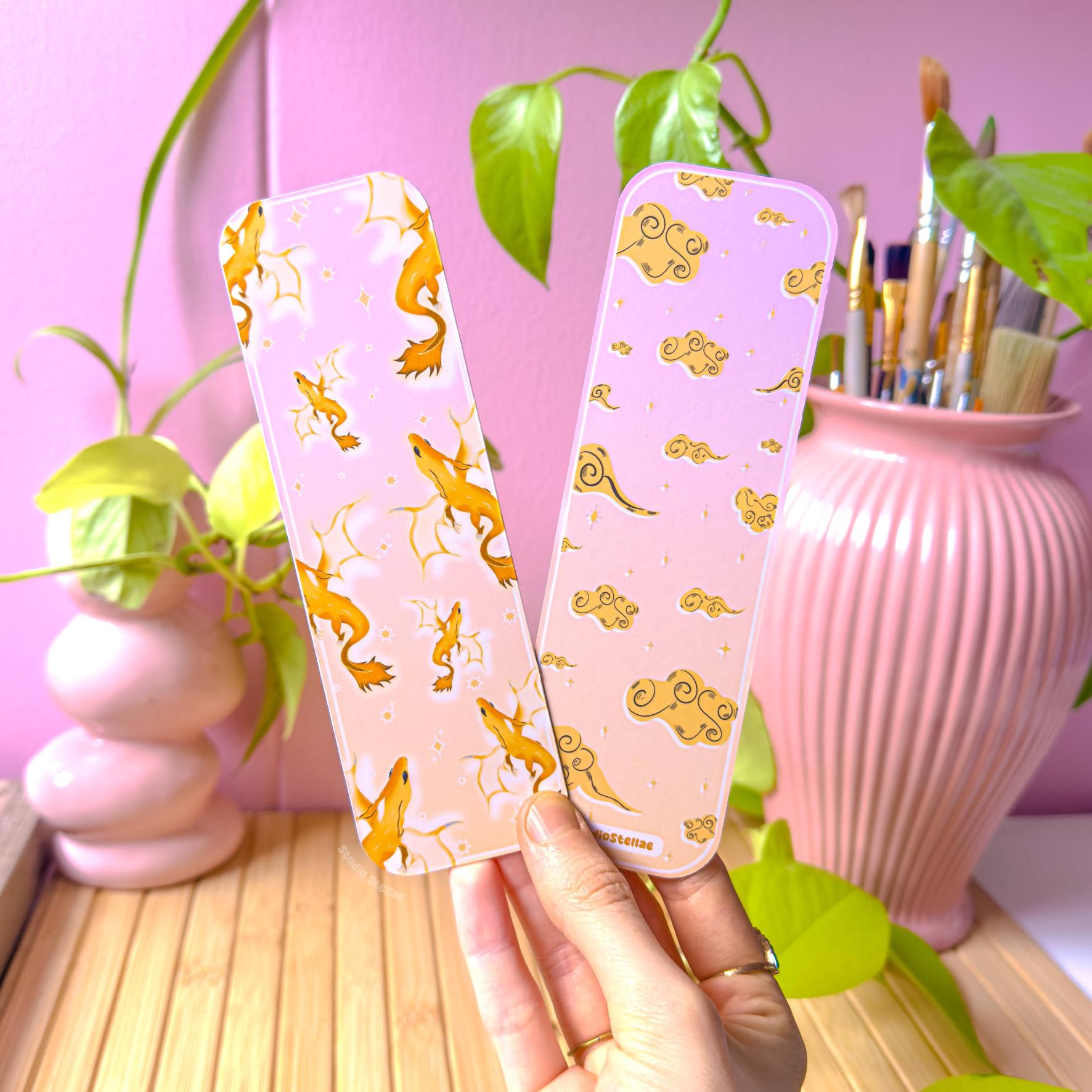 Hand holding the pastel gold dragon bookmark in a cozy workspace with a pink wall, leafy green plant, and a pink ceramic vase filled with paintbrushes in the background. The golden dragon illustration with soft clouds is clearly visible in natural light. This lifestyle image shows real-life scale and how the laminated page marker looks in hand for romantasy readers and fantasy book lovers.