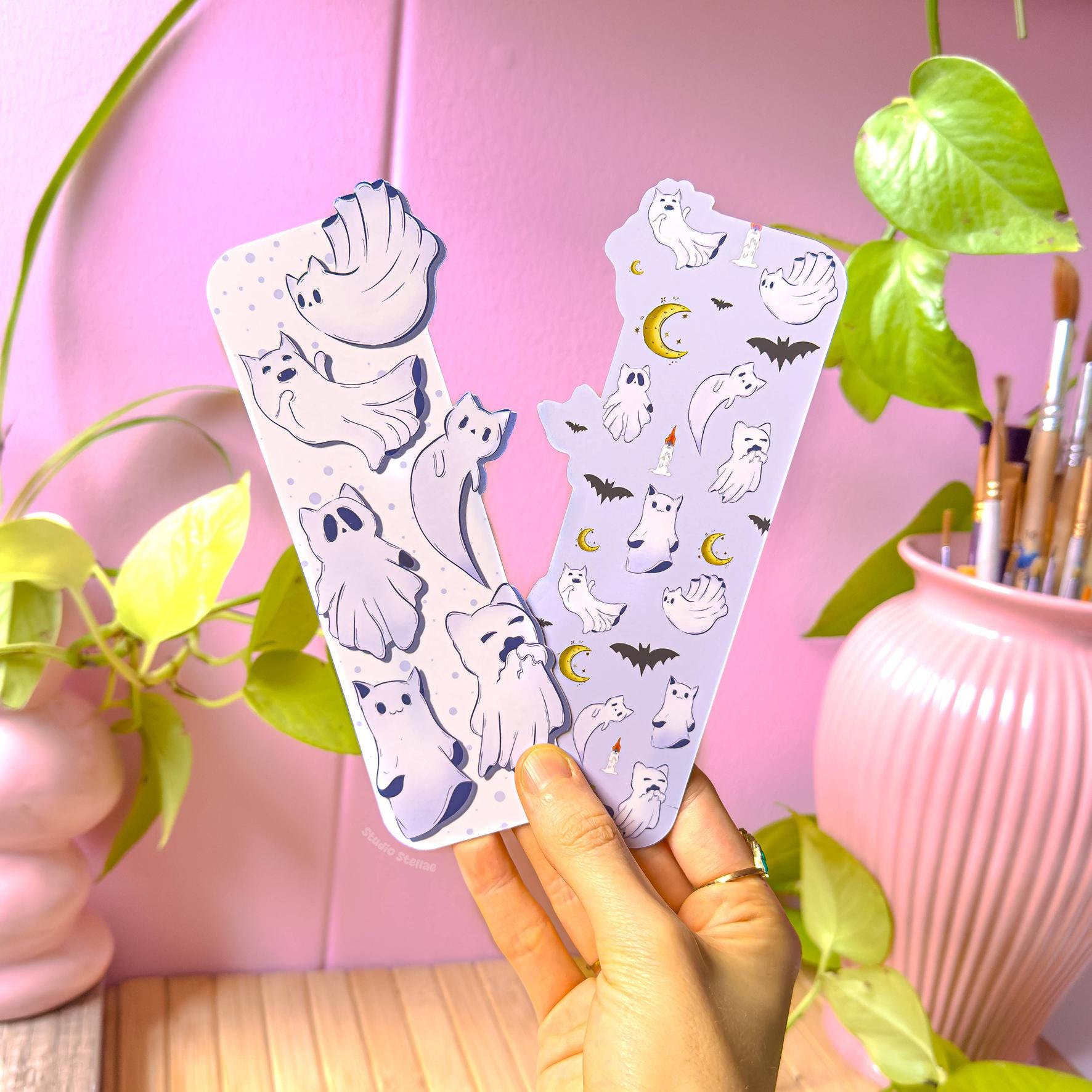 Hand holding two Witchy Cat Ghost Bookmarks in a cozy pastel studio setting with pink wall and green plant in the background. The bookmarks show floating ghost cats, bats, moons, and candles in soft purple tones. This spooky reading accessory is displayed in natural light to highlight the matte lamination and unique die-cut shape, ideal as a Halloween kitty page turner and bookish reader gift.