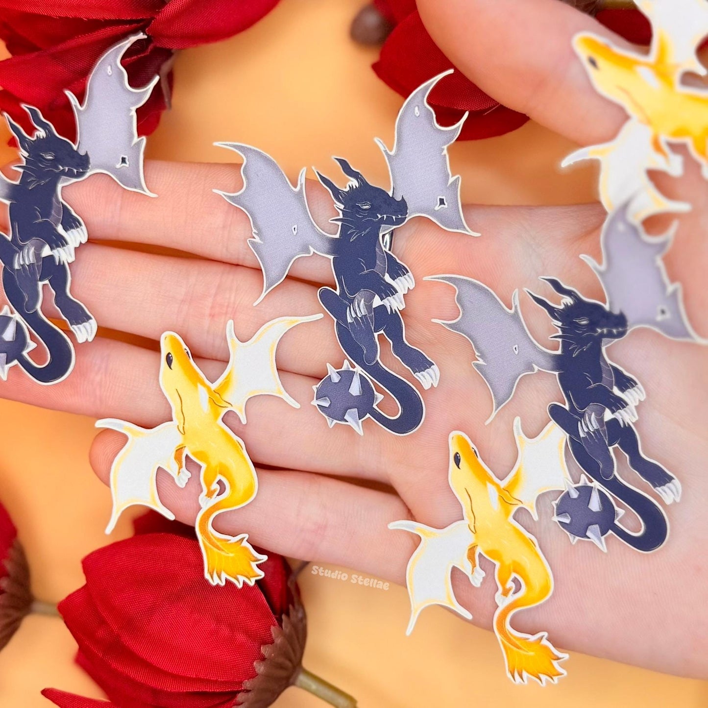 Close-up of the included dragon stickers spread across a hand, showing multiple designs with different poses, shapes, and color tones. The stickers match the bookmark artwork, combining pastel and darker fantasy elements. This detailed shot highlights the bonus items included in the bundle, showing their size, finish, and how they complement the overall set.