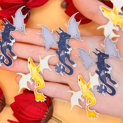 Close-up of the included dragon stickers spread across a hand, showing multiple designs with different poses, shapes, and color tones. The stickers match the bookmark artwork, combining pastel and darker fantasy elements. This detailed shot highlights the bonus items included in the bundle, showing their size, finish, and how they complement the overall set.