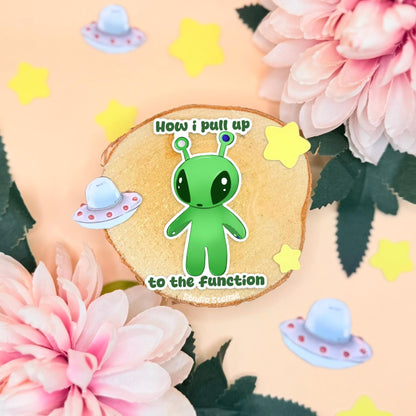 Aftonsparv Alien sticker shown in a soft pastel flat lay with pink flowers and sparkly star charms. The cute green IKEA alien plushie fan art character stands centered with a glossy matte laminated finish and bold die cut shape. This chibi alien sticker features the funny “How I Pull Up to the Function” quote and works perfectly as a laptop sticker, water bottle decal or journal decoration for stationery lovers and alien fans.