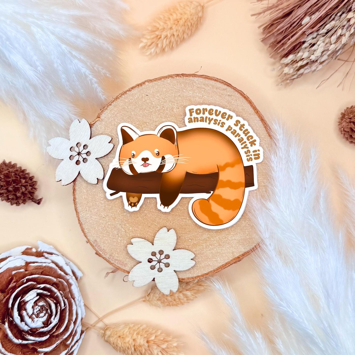 Analysis Paralysis Red Panda sticker photographed in a warm cozy flat lay with wood textures, pinecones and soft neutral tones. The cute kawaii red panda lounges on a tree branch with a tired, overthinking expression, capturing that stuck burnout feeling. This handmade matte laminated vinyl decal features relatable ADHD and neurodivergent humor, perfect as a journal sticker, planner decoration, laptop decal or thoughtful mental health gift.