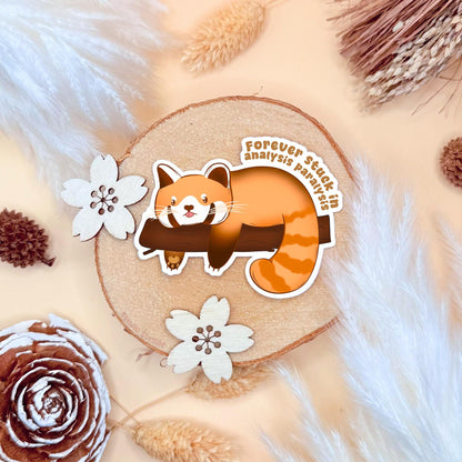 Analysis Paralysis Red Panda sticker photographed in a warm cozy flat lay with wood textures, pinecones and soft neutral tones. The cute kawaii red panda lounges on a tree branch with a tired, overthinking expression, capturing that stuck burnout feeling. This handmade matte laminated vinyl decal features relatable ADHD and neurodivergent humor, perfect as a journal sticker, planner decoration, laptop decal or thoughtful mental health gift.