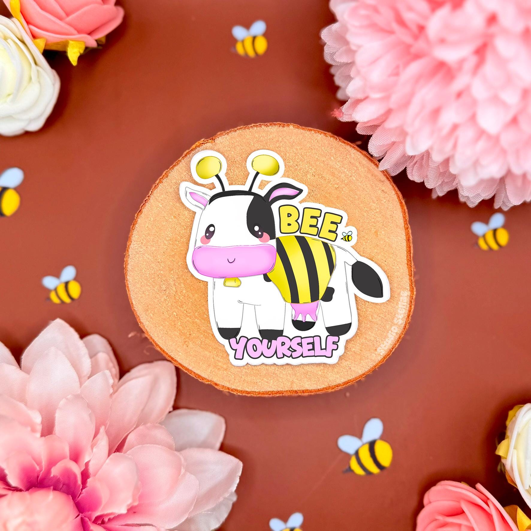 Bee Yourself cow sticker styled in a soft pastel flat lay with pink flowers and warm cork textures. The kawaii cow wears a bumblebee costume with tiny wings and stripes, paired with the positive affirmation quote “Bee Yourself.” This handmade matte laminated vinyl decal combines cute animal art with LGBTQ+ pride and neurodivergent positivity, perfect for decorating laptops, water bottles, planners, journals and colorful stationery gifts.