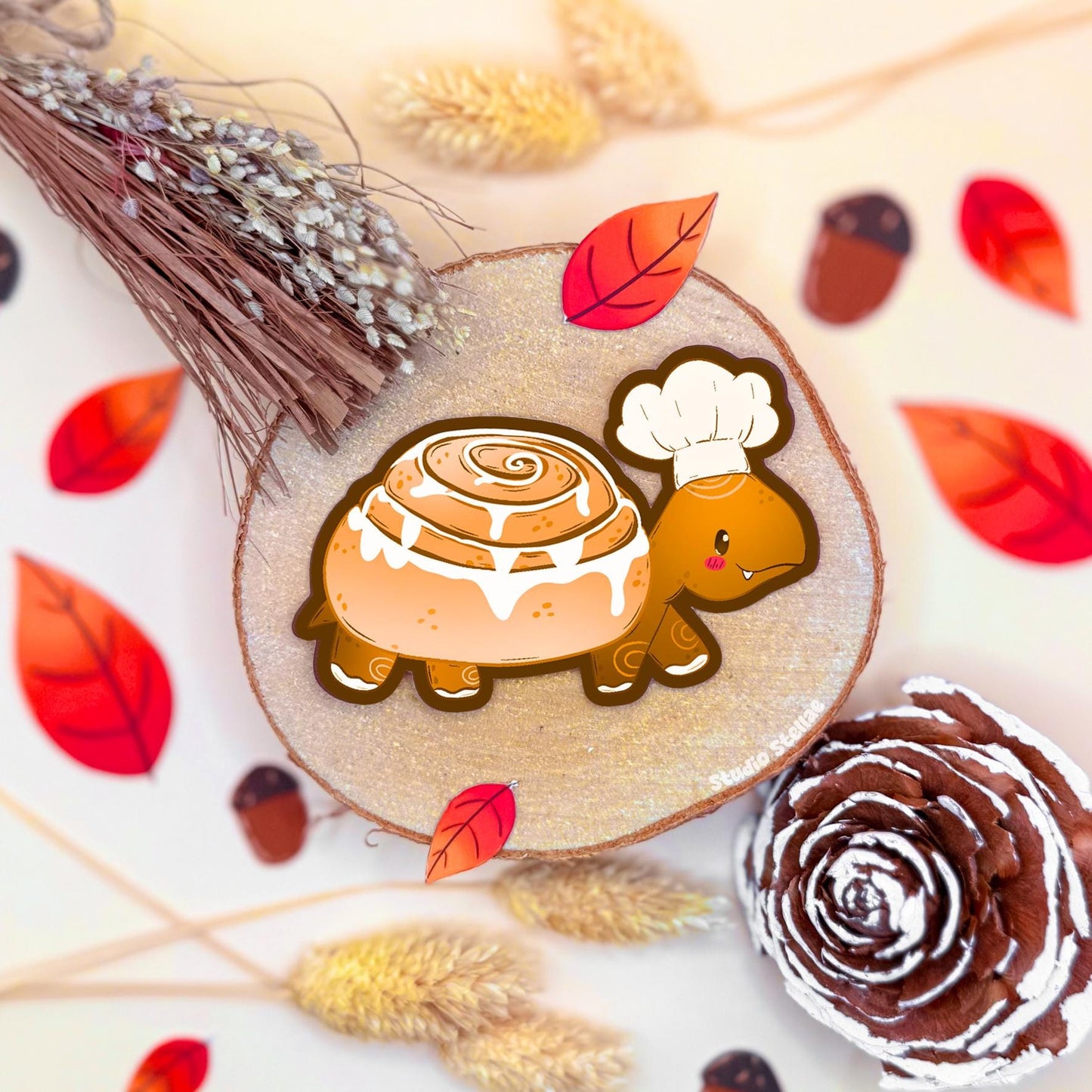 Cinnamon Roll Turtle sticker styled in a warm cozy flat lay with dried wheat, autumn leaves and bakery inspired textures. The cute turtle shaped like a frosted cinnamon roll rests in the center, combining sweet pastry art with soft kawaii animal charm. This handmade matte vinyl sticker has a smooth finish and bold die cut outline, perfect for decorating journals, planners, laptops and gifts for comfort food and bakery lovers.