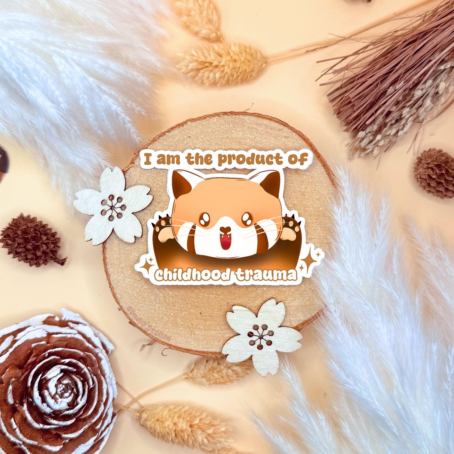 Cute red panda chibi sticker styled in a warm cozy flat lay with dried wheat and soft neutral textures. The kawaii red panda lies cutely with the funny dark humor quote “I am the product of childhood trauma,” blending adorable cartoon art with relatable therapy humor. This handmade matte laminated vinyl decal adds personality to laptops, planners, journals and water bottles as a playful mental health joke.