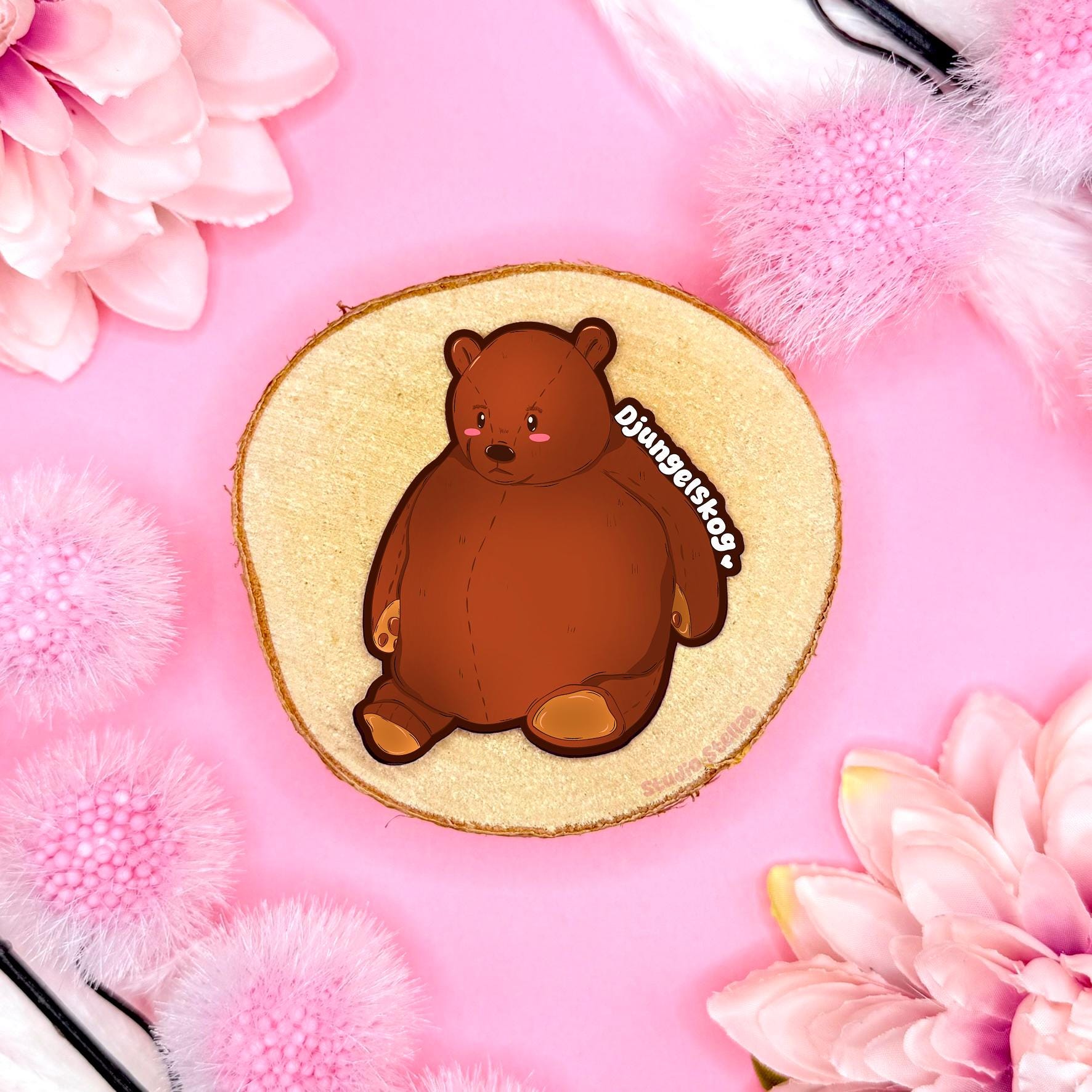 Djungelskog bear sticker styled in a soft pastel flat lay with pink flowers and warm cork textures. The cute chubby brown teddy bear is illustrated as cozy IKEA plushie fan art with a round cuddly shape and gentle expression. This handmade matte laminated vinyl decal captures the comforting stuffed animal vibe, perfect for decorating journals, planners, laptops and water bottles or gifting to plushie and bear lovers.