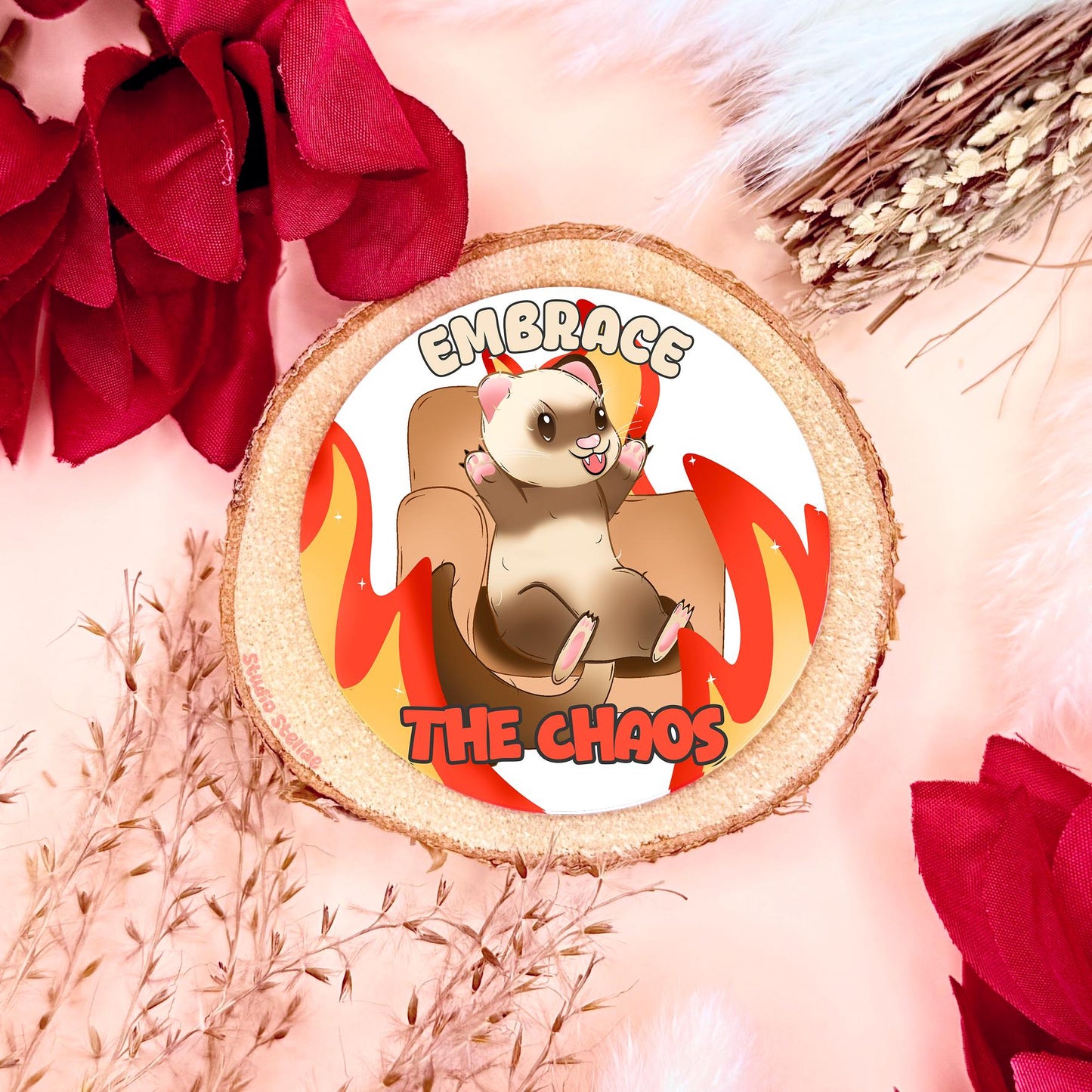 Embrace the Chaos ferret sticker styled in a warm flat lay with red flowers and cork textures. The cute kawaii ferret sits on a chair surrounded by cartoon flames with the phrase “Embrace the Chaos,” blending adorable animal art with funny chaotic humor. This handmade matte laminated vinyl decal adds playful burnout energy to laptops, planners, journals and water bottles as a relatable mental health meme.