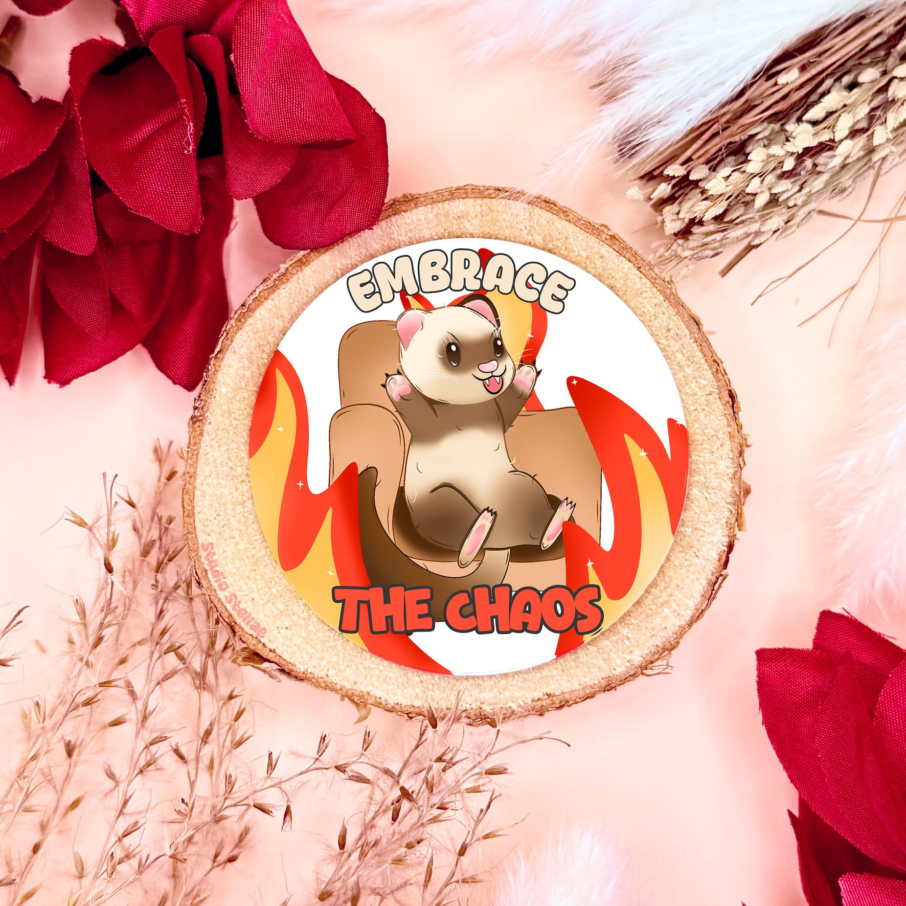 Embrace the Chaos ferret sticker styled in a warm flat lay with red flowers and cork textures. The cute kawaii ferret sits on a chair surrounded by cartoon flames with the phrase “Embrace the Chaos,” blending adorable animal art with funny chaotic humor. This handmade matte laminated vinyl decal adds playful burnout energy to laptops, planners, journals and water bottles as a relatable mental health meme.