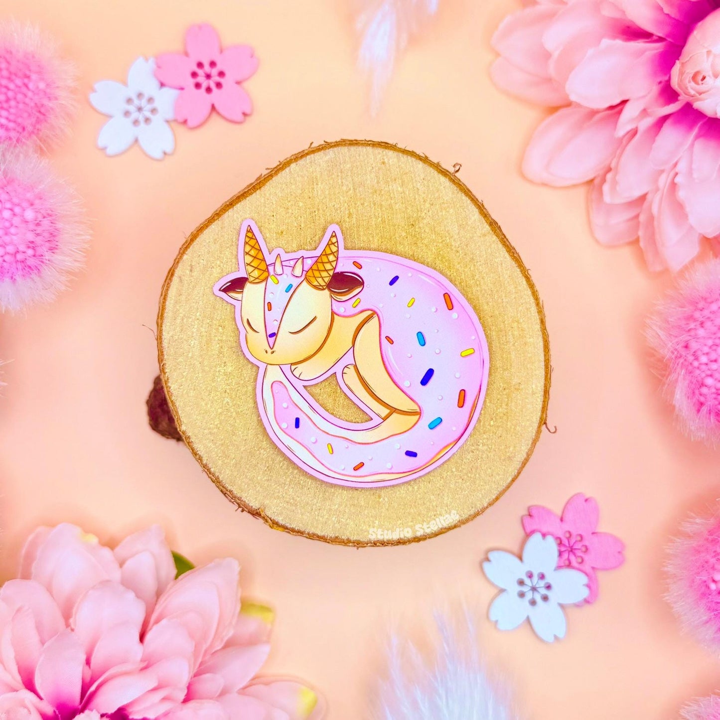 Cute dragon sticker styled in a soft pastel flat lay with pink flowers and warm cork textures. The tiny frosted sugar cookie dragon curls up in pink icing like a pastry, blending kawaii dragon art with cozy fantasy bakery vibes. This handmade matte laminated vinyl decal looks sweet and magical, perfect for decorating journals, planners, laptops and water bottles or gifting to dessert and stationery lovers.