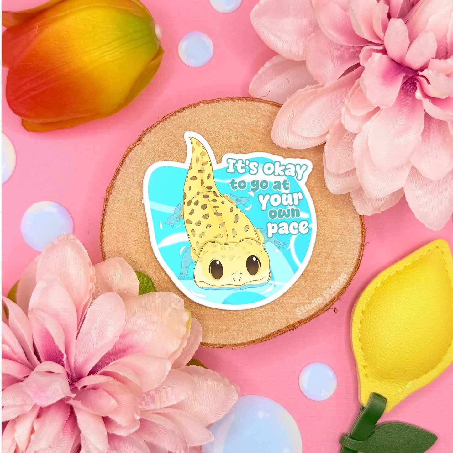 Cute gecko sticker styled in a soft pastel flat lay with pink flowers and warm cork textures. The sweet yellow lizard sits on a blue heart with the quote “It’s okay to go at your own pace,” creating a gentle motivational affirmation. This handmade matte laminated vinyl decal blends kawaii reptile art with cozy self care vibes, perfect for decorating planners, journals, laptops and water bottles or gifting to reptile lovers.