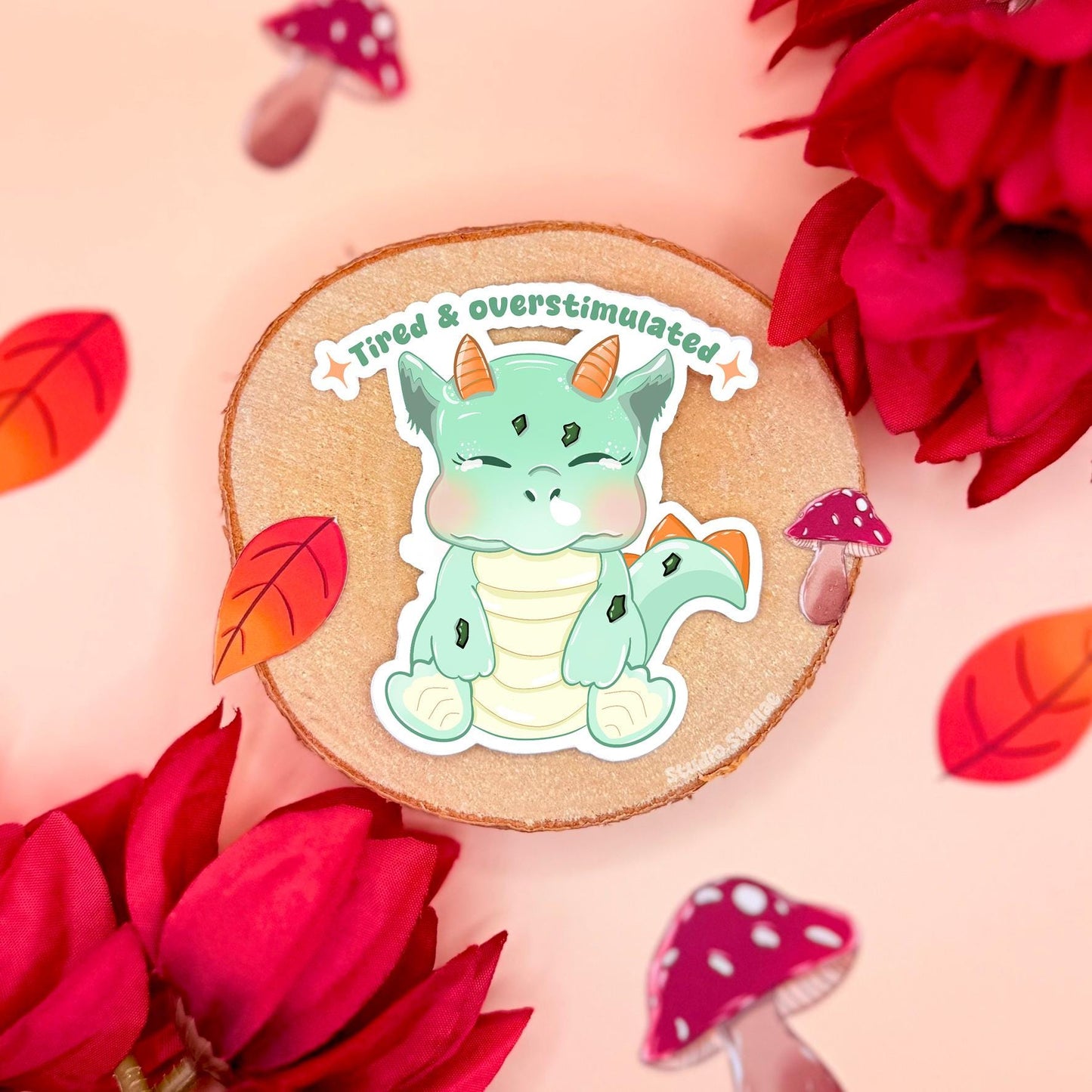 Tired and overstimulated baby dinosaur sticker styled in a soft pastel flat lay with cozy textures and cork background. The cute kawaii dino looks exhausted and slumped over, capturing relatable burnout and neurodivergent overwhelm humor. This handmade matte laminated vinyl decal blends gentle mental health art with adorable dinosaur charm, perfect for decorating planners, journals, laptops and water bottles.