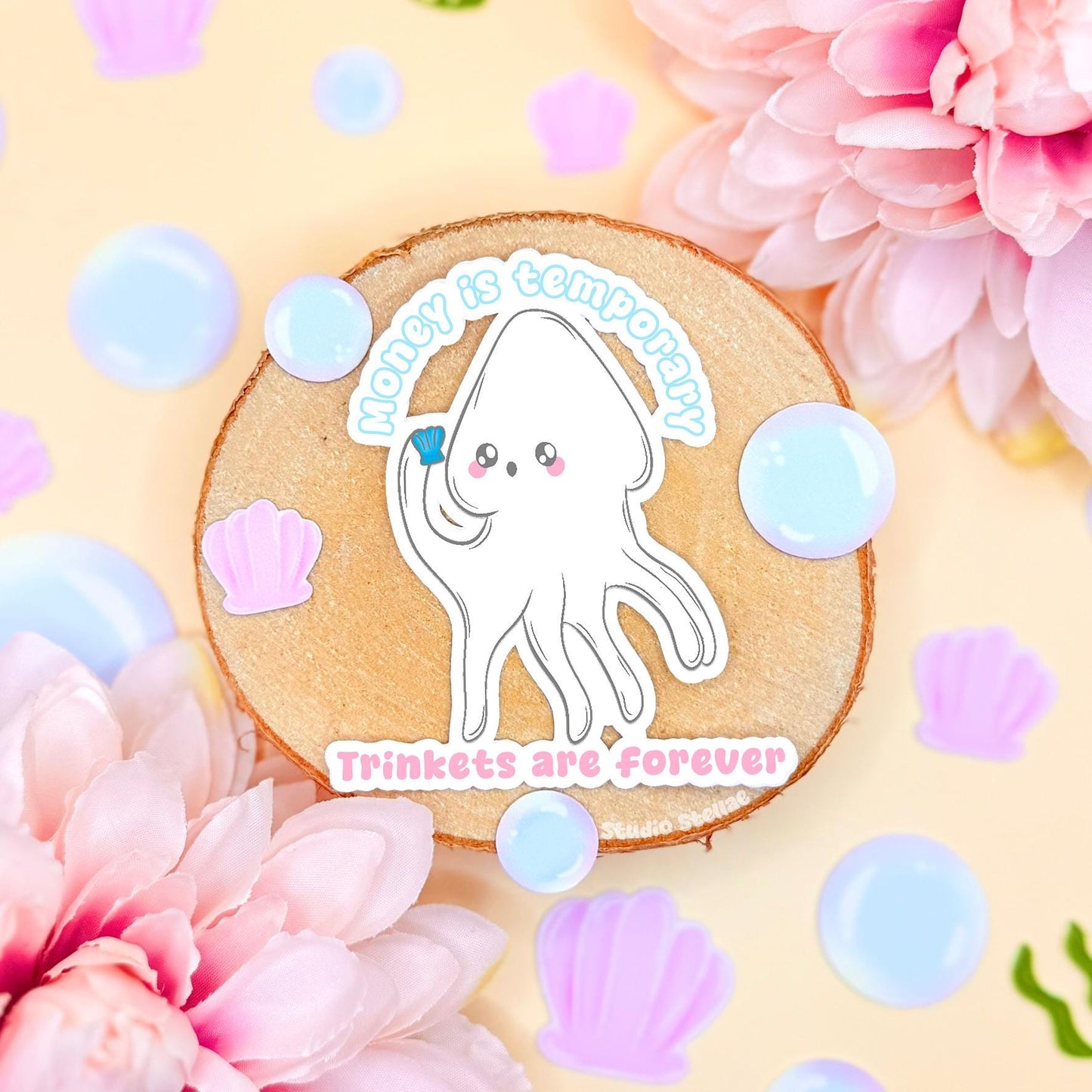 White squid sticker styled in a soft pastel flat lay with flowers and cozy textures on a cork background. The cute kawaii ocean creature includes the funny quote “Money is temporary, trinkets are forever,” blending adorable sea life art with playful anti-capitalist humor. This handmade matte laminated vinyl decal is perfect for decorating laptops, planners, journals and water bottles as a trinket lover gift.