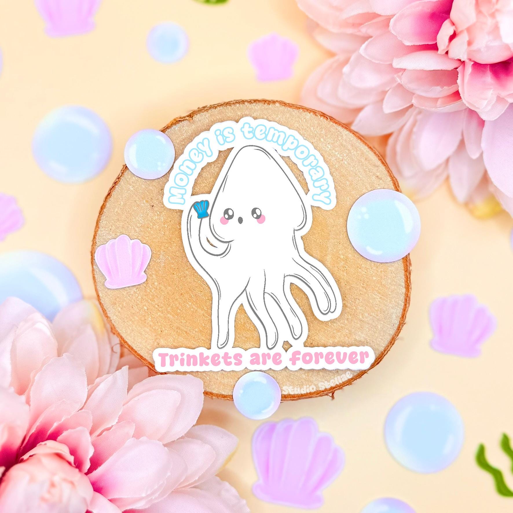 White squid sticker styled in a soft pastel flat lay with flowers and cozy textures on a cork background. The cute kawaii ocean creature includes the funny quote “Money is temporary, trinkets are forever,” blending adorable sea life art with playful anti-capitalist humor. This handmade matte laminated vinyl decal is perfect for decorating laptops, planners, journals and water bottles as a trinket lover gift.