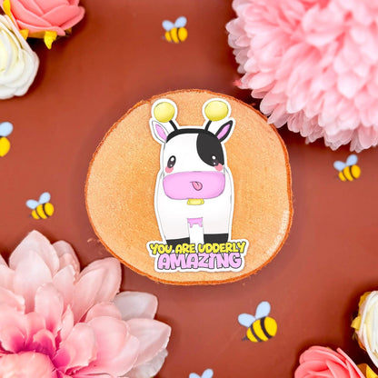 Cow Bumblebee sticker displayed in a soft pastel flat lay with pink flowers and warm cork textures. The cute chibi cow wears a yellow bee costume with tiny wings and stripes beside the affirmation quote “You Are Udderly Amazing.” This handmade matte laminated vinyl decal blends kawaii animal art with feel good positivity, perfect for decorating journals, planners, laptops and water bottles or gifting to a friend who needs encouragement.