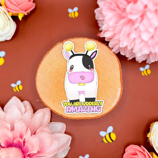 Cow Bumblebee sticker displayed in a soft pastel flat lay with pink flowers and warm cork textures. The cute chibi cow wears a yellow bee costume with tiny wings and stripes beside the affirmation quote “You Are Udderly Amazing.” This handmade matte laminated vinyl decal blends kawaii animal art with feel good positivity, perfect for decorating journals, planners, laptops and water bottles or gifting to a friend who needs encouragement.