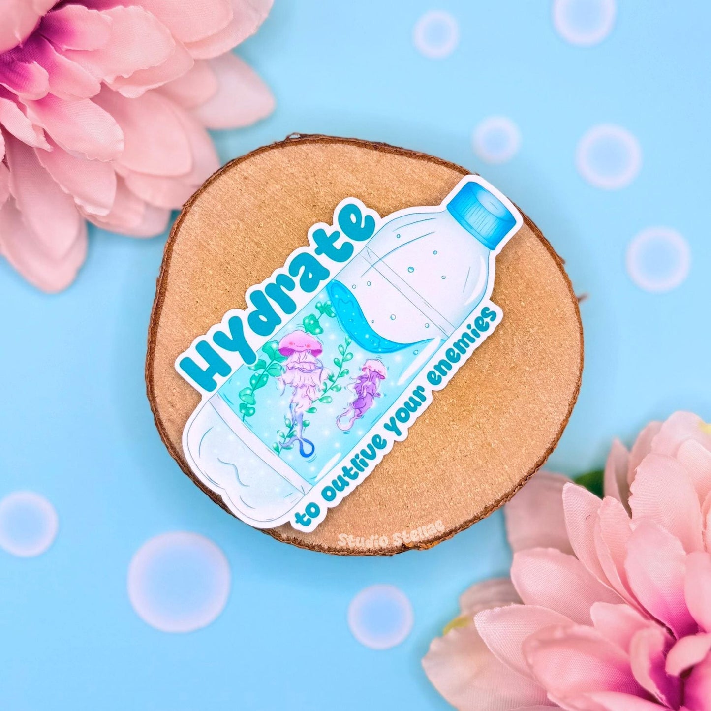 Cute jellyfish water bottle sticker styled in a soft pastel flat lay with pink flowers and warm cork textures. A sparkly blue moon jellyfish floats beside the funny quote “Hydrate to outlive your enemies,” blending adorable ocean animal art with sarcastic self care humor. This handmade matte laminated vinyl decal is perfect for decorating water bottles, planners, laptops and journals as a playful hydration reminder.