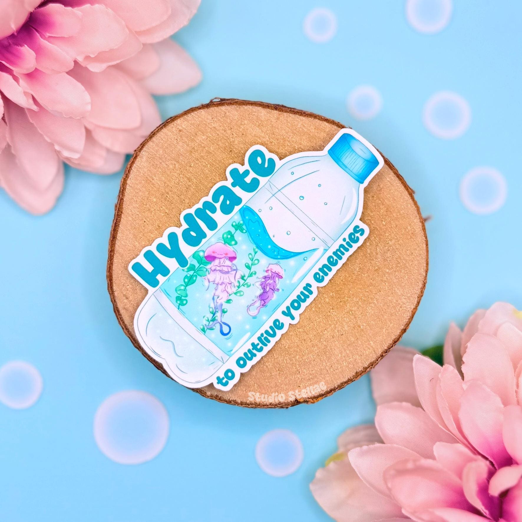 Cute jellyfish water bottle sticker styled in a soft pastel flat lay with pink flowers and warm cork textures. A sparkly blue moon jellyfish floats beside the funny quote “Hydrate to outlive your enemies,” blending adorable ocean animal art with sarcastic self care humor. This handmade matte laminated vinyl decal is perfect for decorating water bottles, planners, laptops and journals as a playful hydration reminder.