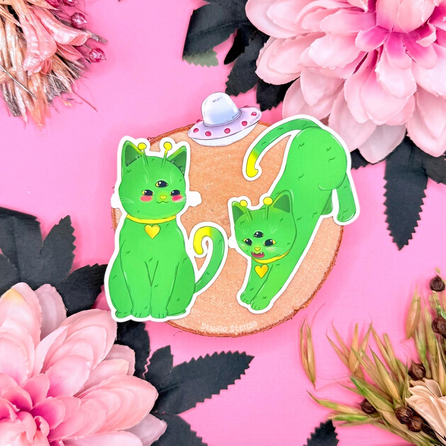 Alien cat sticker set displayed on a soft pink background with flowers and decorative props, featuring three green cosmic kitties with playful expressions and sci fi details. This kawaii space cat sticker pack highlights the chibi alien cat designs and matte finish, showing a cute outer space themed sticker set perfect for decorating laptops, planners, journals, or water bottles and gifting to cat lovers who enjoy cosmic aesthetics and quirky fantasy companions.