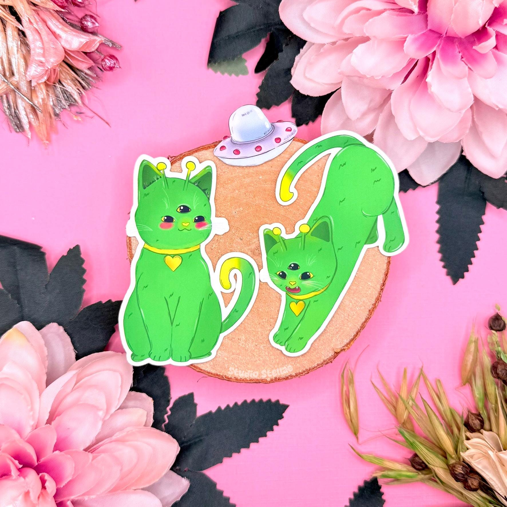 Alien cat sticker set displayed on a soft pink background with flowers and decorative props, featuring three green cosmic kitties with playful expressions and sci fi details. This kawaii space cat sticker pack highlights the chibi alien cat designs and matte finish, showing a cute outer space themed sticker set perfect for decorating laptops, planners, journals, or water bottles and gifting to cat lovers who enjoy cosmic aesthetics and quirky fantasy companions.