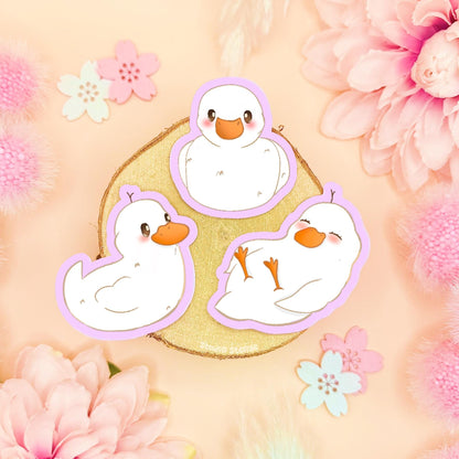 Baby duck sticker set featuring three tiny pastel duckling stickers arranged on a soft pink background with flowers and decorative props. This kawaii duck sticker pack shows cute white duck characters in different poses, highlighting their matte finish and soft colors. Handmade waterproof baby duck decals perfect for decorating laptops, planners, journals, scrapbooks, or water bottles, and a sweet gift for duck lovers, stationery fans, and anyone who loves cozy pastel animal stickers.