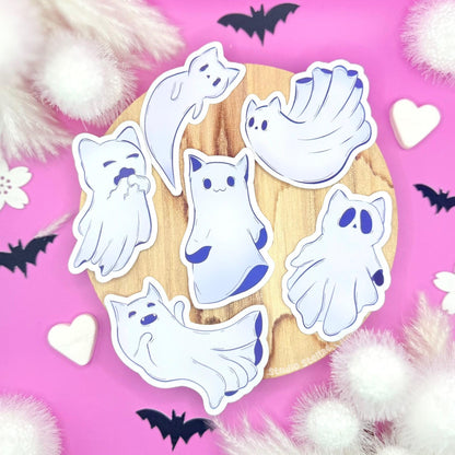 Full Ghost Kitty sticker set displayed on a soft pastel pink background scattered with tiny hearts, creating a cute yet slightly spooky pastel goth aesthetic. Six kawaii ghost cats float together, each with its own playful, sleepy, or mischievous expression. This main image shows the entire Halloween sticker pack at a glance while highlighting clean die-cut edges, vibrant pastel colors, and a smooth matte laminated finish. Perfect for planners, journals, laptops, and cozy seasonal Sticker lovers