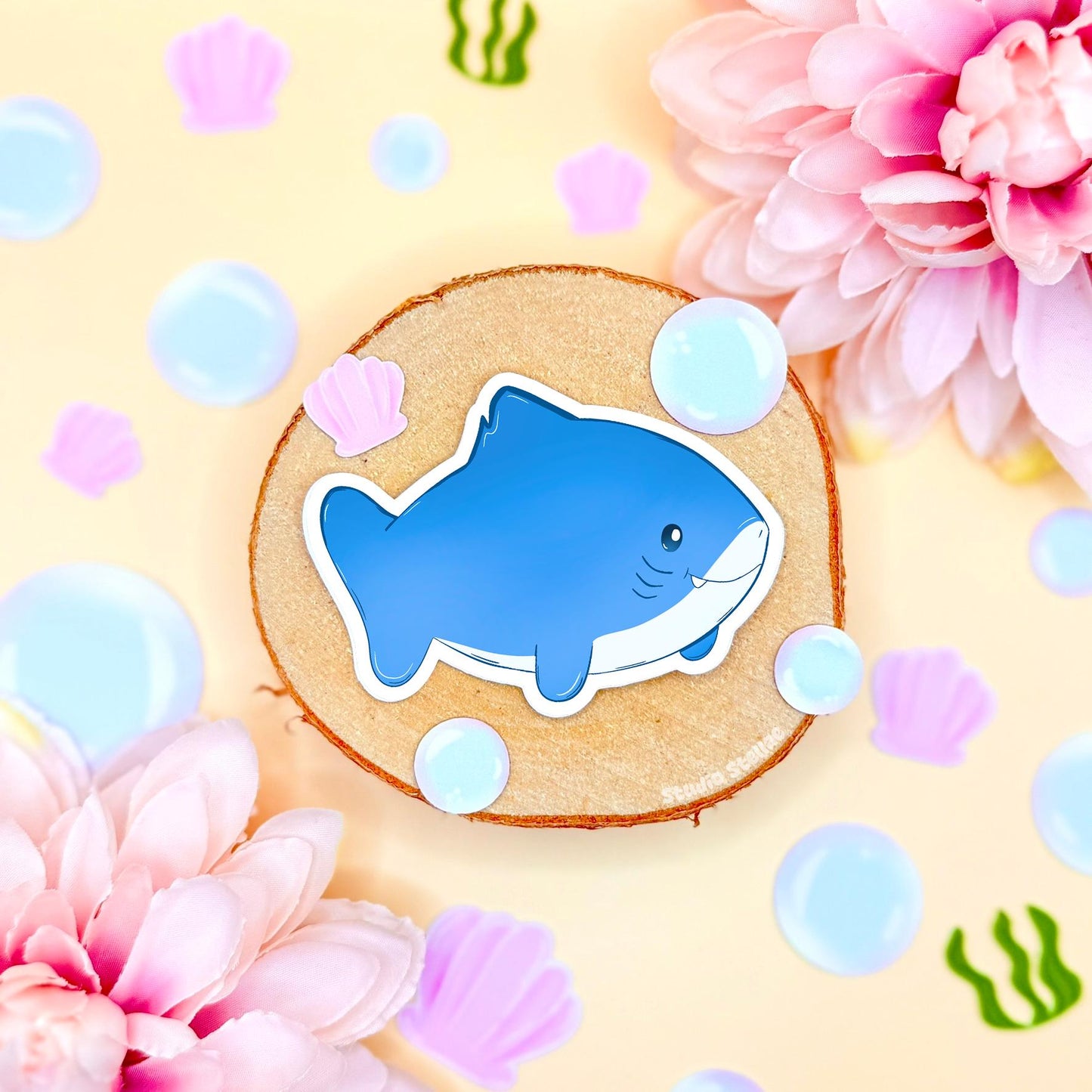 Close-up of a blue chibi shark sticker on a cork background with bubbles and pastel decor, showing its rounded body, tiny fins and friendly smile. The smooth matte laminate gives a soft premium finish while protecting against scratches and moisture. This cute shark decal adds fun ocean vibes to water bottles, planners, journals and laptops and is part of the colorful kawaii sea life sticker pack.