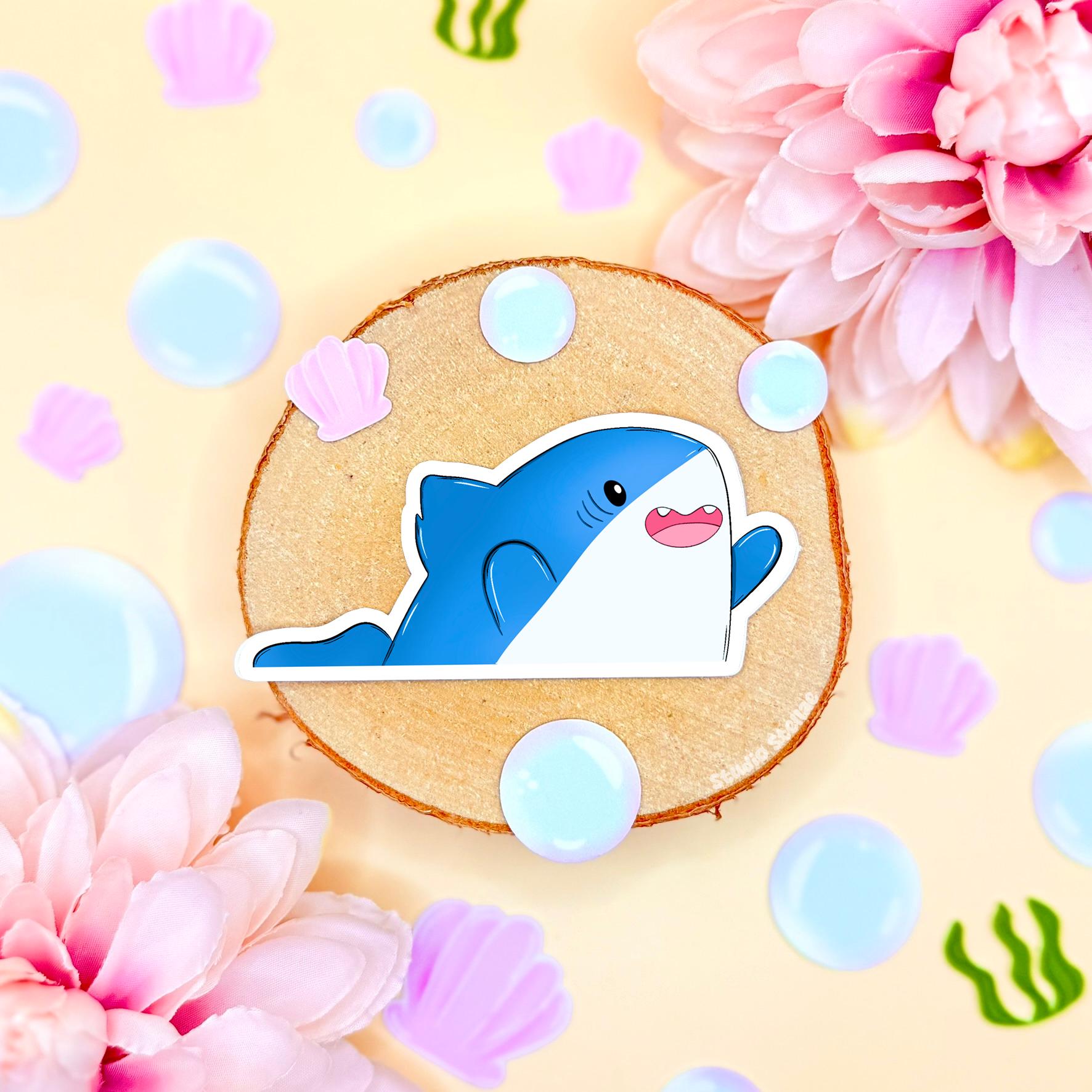 Close-up of a cheerful blue chibi shark sticker with tiny fins raised like it’s celebrating, photographed against a soft pastel cork background with bubbles and flowers. The happy expression and playful pose give this kawaii ocean animal decal extra personality and charm. Matte laminated, waterproof and scratch resistant, this cute shark sticker is perfect for decorating laptops, planners, journals and water bottles, adding fun sea life vibes to any cozy stationery setup.