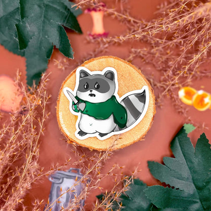 Detailed view of a rogue raccoon sticker posed with fantasy gear on a styled tabletop, emphasizing costume elements, soft shading, and handmade illustration style. This image showcases the character personality within the DnD raccoon sticker set and helps customers see scale and finish. Perfect for tabletop RPG fans looking to decorate gaming accessories or planners with cute fantasy animal art.