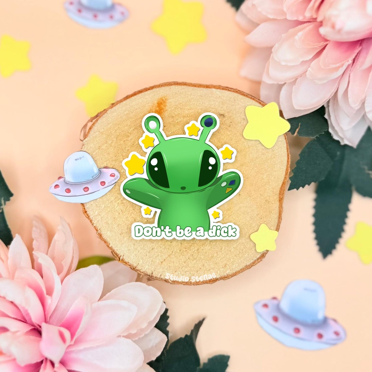 A handmade Aftonsparv Alien Sticker styled as cute IKEA plushie fan art, photographed on a soft pastel pink floral background. The small green chibi alien has sparkly stars and a bold die cut shape, shown as a matte laminated vinyl decal for laptops, water bottles, notebooks and journals. This Don’t Be a Dick funny quote sticker blends kawaii cute art with humor, perfect for stationery lovers and small business handmade gifts.