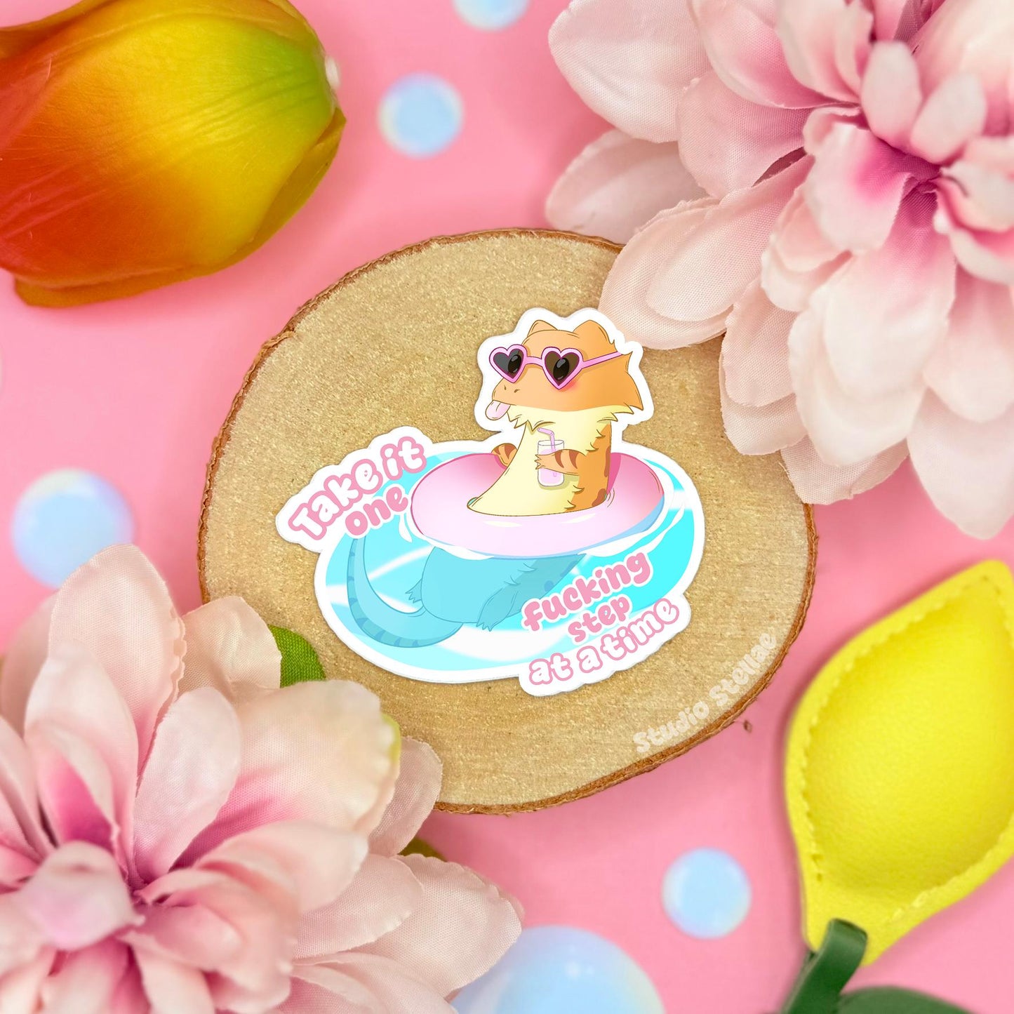 Cute bearded dragon sticker styled in a soft pastel flat lay with pink flowers and warm cork textures. The smiling pink lizard floats on a blue puddle beside the quote “Take it one fcking step at a time,” mixing gentle self care with funny sarcastic humor. This handmade matte vinyl decal works perfectly as a mental health reminder on laptops, water bottles, planners and journals for reptile lovers.