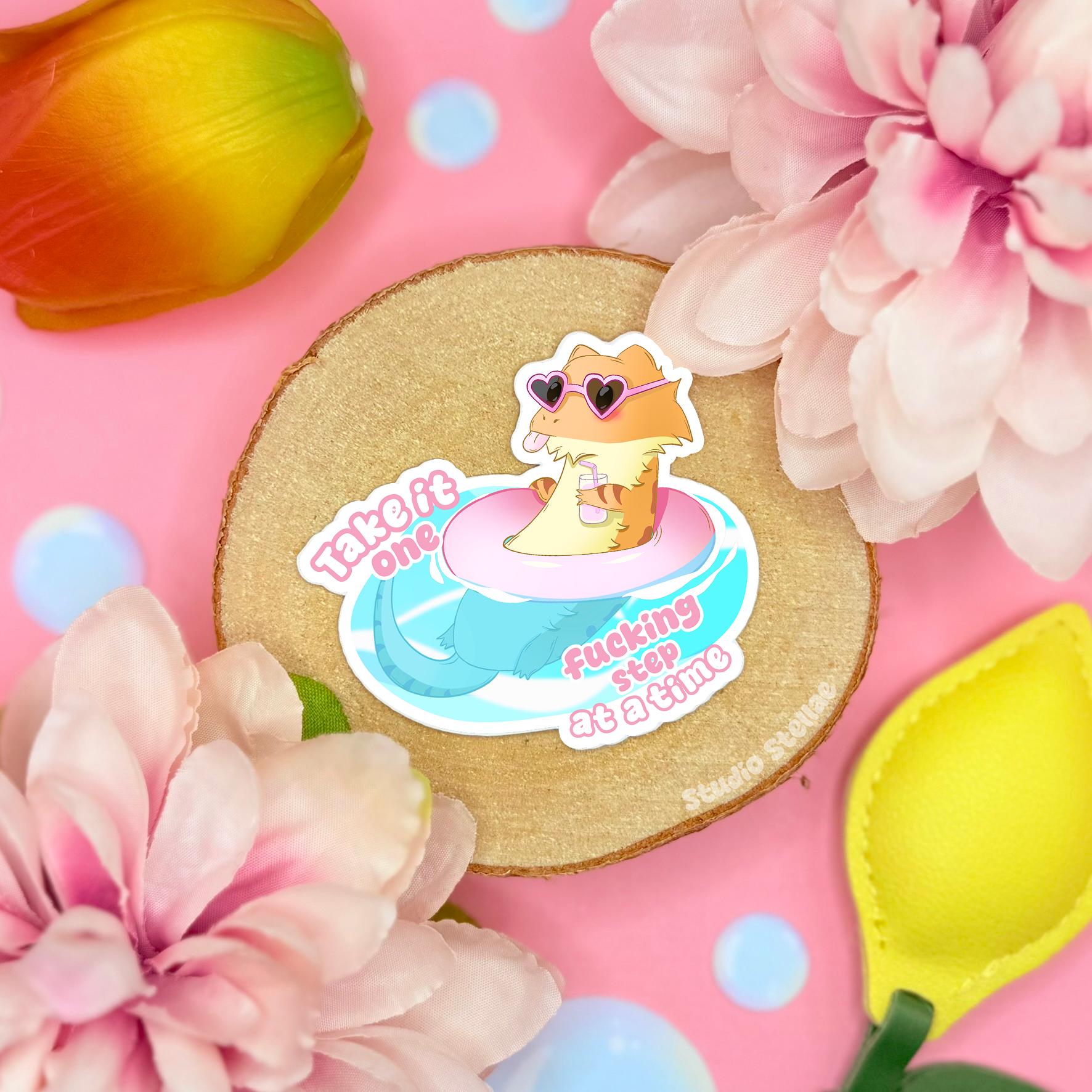 Cute bearded dragon sticker styled in a soft pastel flat lay with pink flowers and warm cork textures. The smiling pink lizard floats on a blue puddle beside the quote “Take it one fcking step at a time,” mixing gentle self care with funny sarcastic humor. This handmade matte vinyl decal works perfectly as a mental health reminder on laptops, water bottles, planners and journals for reptile lovers.