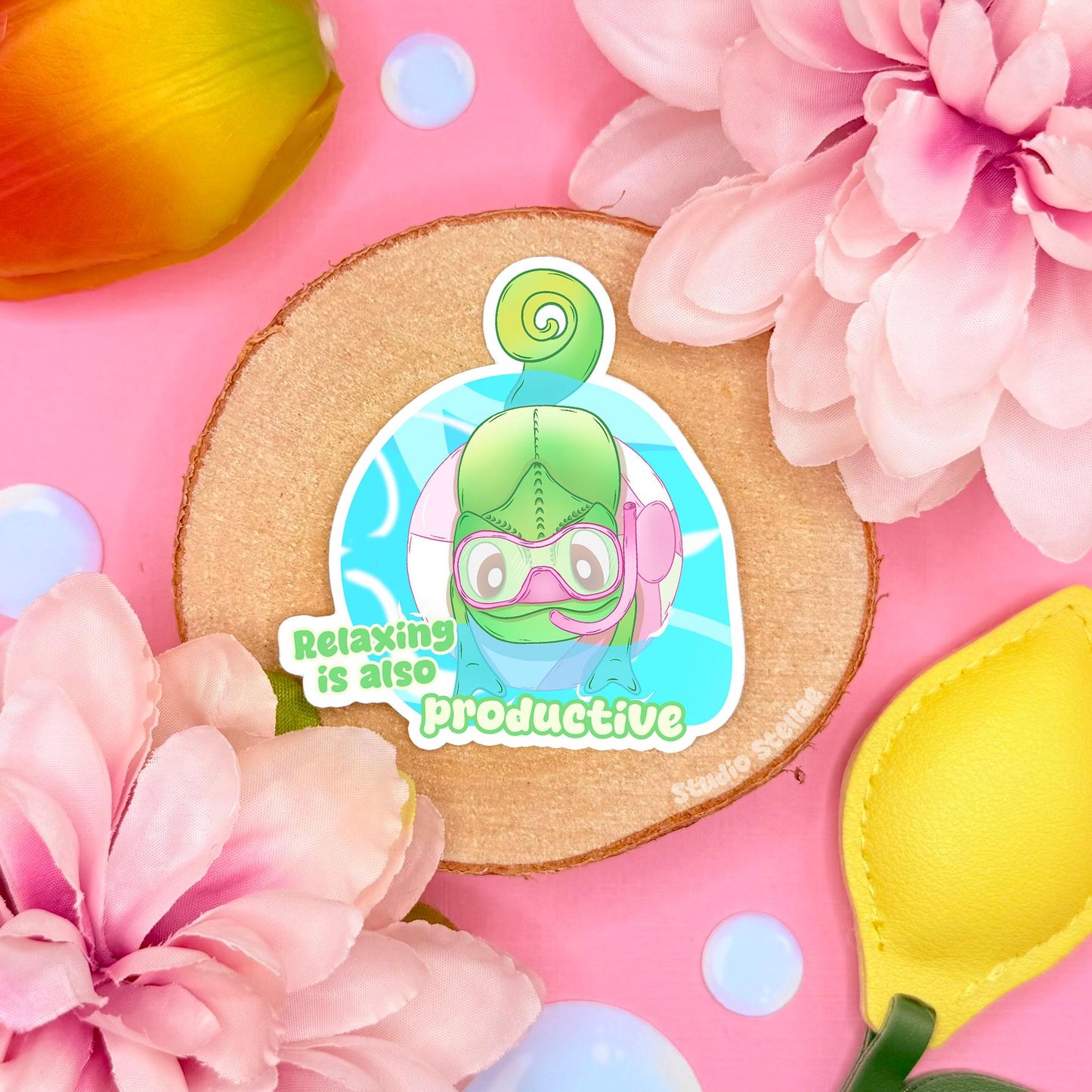 Cute chameleon sticker displayed in a soft pastel flat lay with pink flowers and warm cork textures. The green kawaii lizard curls up peacefully beside the quote “Relaxing is also productive,” creating a gentle mental health and self care reminder. This handmade matte laminated vinyl decal adds calming positivity to journals, planners, laptops and water bottles, making it a sweet motivational gift for reptile lovers.