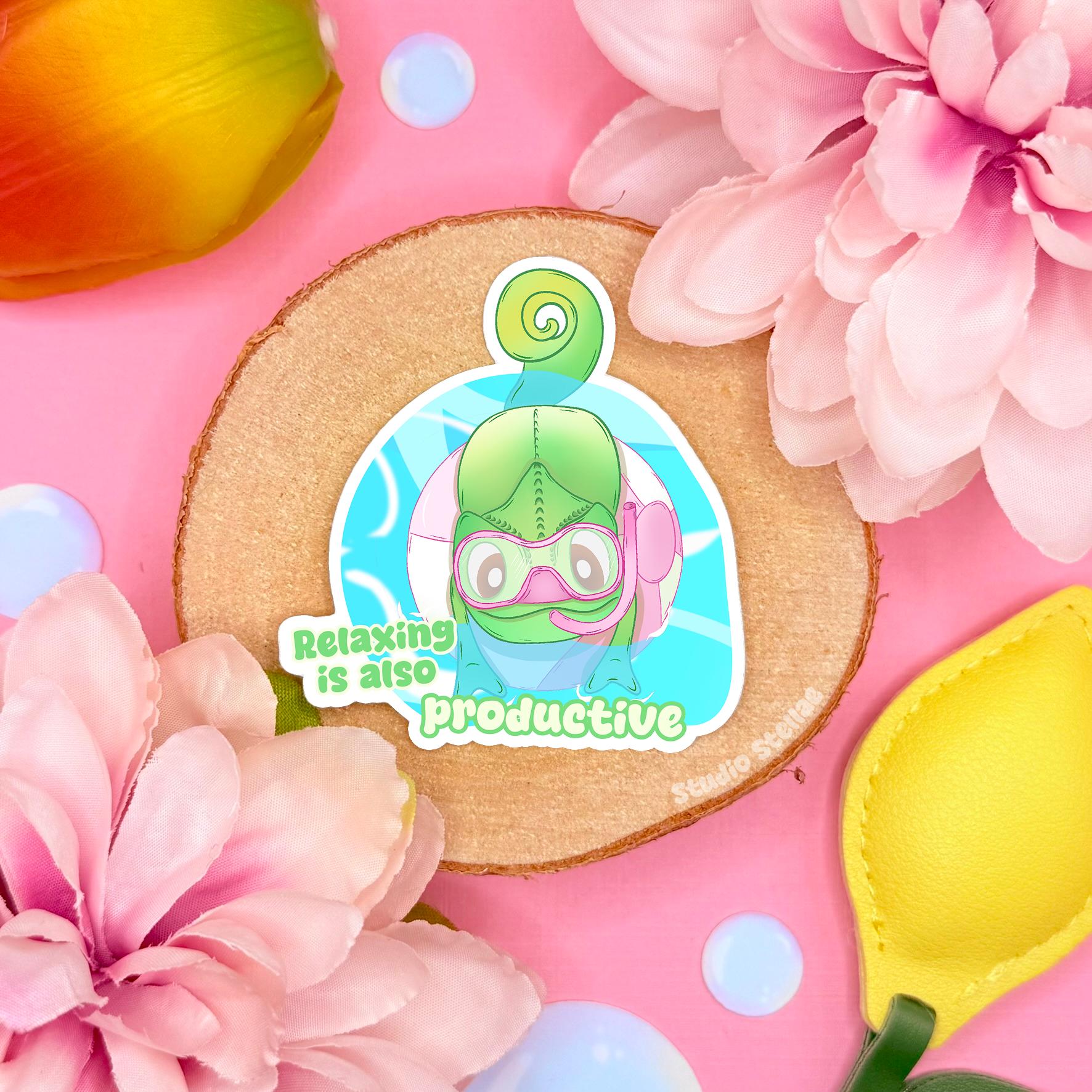 Cute chameleon sticker displayed in a soft pastel flat lay with pink flowers and warm cork textures. The green kawaii lizard curls up peacefully beside the quote “Relaxing is also productive,” creating a gentle mental health and self care reminder. This handmade matte laminated vinyl decal adds calming positivity to journals, planners, laptops and water bottles, making it a sweet motivational gift for reptile lovers.