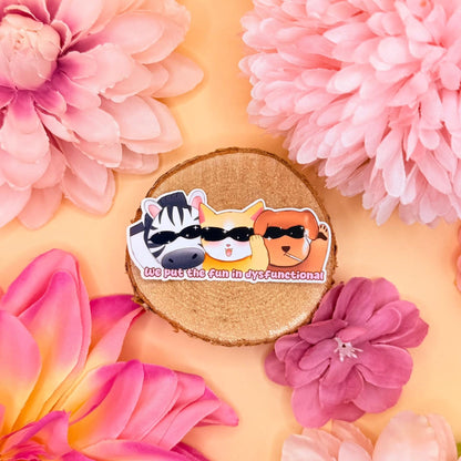Colorful Animal Party sticker styled in a soft pastel flat lay with pink flowers and warm cork textures. The chibi cat, dog and zebra characters stand together with the funny quote “We put the fun in dysfunctional,” mixing cute animal art with dark humor. This handmade matte laminated vinyl decal adds a playful yet relatable mental health joke, perfect for decorating laptops, planners, journals and water bottles.