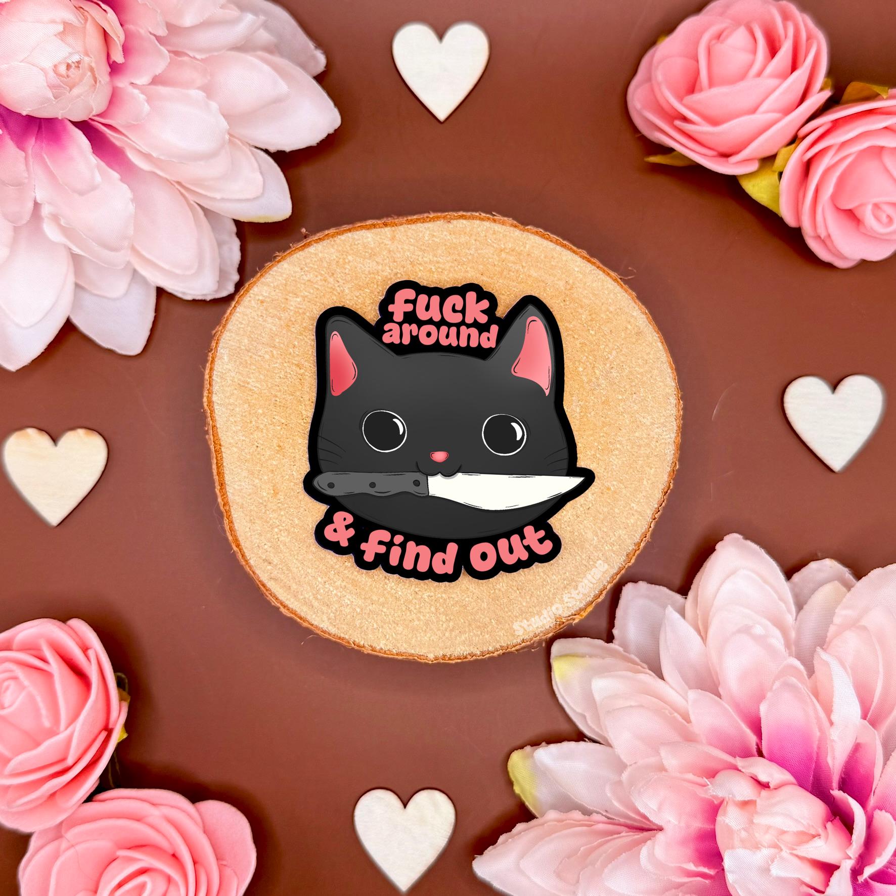 Funny black cat meme sticker styled in a soft flat lay with pink flowers and warm cork textures. The sassy chibi kitty stares with attitude beside the bold “fuck around & find out” FAFO quote, blending cute kawaii cat art with sarcastic adult humor. This handmade matte laminated vinyl decal adds chaotic gremlin energy to laptops, planners, notebooks and water bottles as a playful warning sticker.