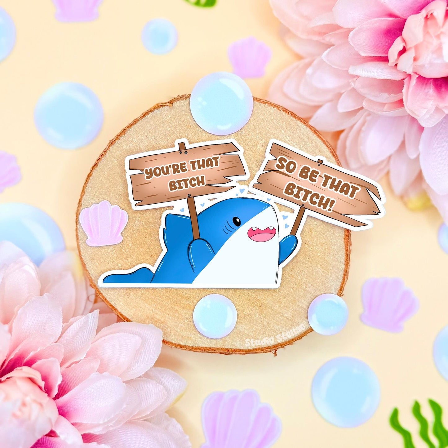 Cute shark sticker styled in a soft pastel flat lay with pink flowers and warm cork textures. The chibi blue shark holds a sign that reads “You’re that bitch,” creating a funny motivational meme with bold confidence energy. This handmade matte laminated vinyl decal blends cute animal art with self love humor, perfect for decorating laptops, planners, notebooks and water bottles as a daily hype reminder.