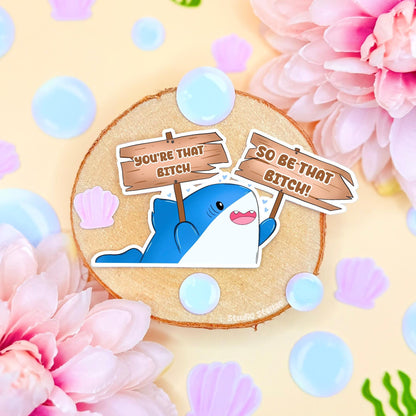 Cute shark sticker styled in a soft pastel flat lay with pink flowers and warm cork textures. The chibi blue shark holds a sign that reads “You’re that bitch,” creating a funny motivational meme with bold confidence energy. This handmade matte laminated vinyl decal blends cute animal art with self love humor, perfect for decorating laptops, planners, notebooks and water bottles as a daily hype reminder.