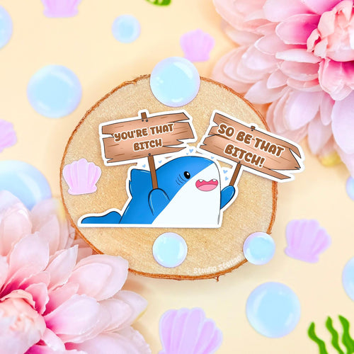 Cute shark sticker styled in a soft pastel flat lay with pink flowers and warm cork textures. The chibi blue shark holds a sign that reads “You’re that bitch,” creating a funny motivational meme with bold confidence energy. This handmade matte laminated vinyl decal blends cute animal art with self love humor, perfect for decorating laptops, planners, notebooks and water bottles as a daily hype reminder.