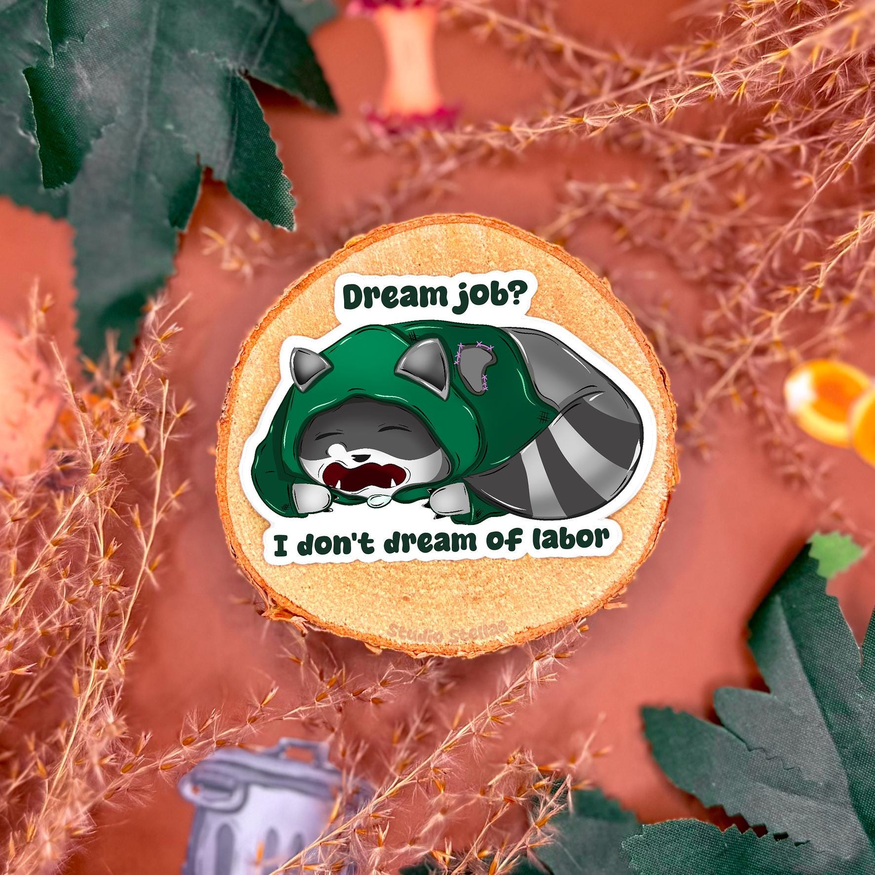 Cute rogue raccoon sticker styled in a warm autumn flat lay with leaves and cork textures. The sleepy kawaii trash panda lies flat with tired eyes beside the quote “Dream Job? I Don’t Dream of Labor,” capturing relatable anti-work and burnout humor. This handmade matte laminated vinyl decal blends soft animal art with sarcastic rebellion, perfect for decorating laptops, planners and water bottles.